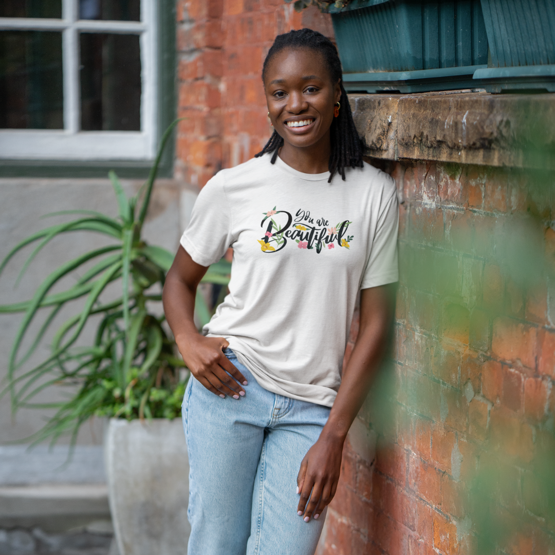 Woman wearing a 'She is Beautiful' t-shirt standing against a brick wall.